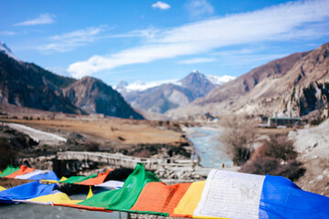 Colorful prayer flags flutter in the wind as a river winds through the rugged valleys of the Annapurna Circuit. The majestic snow-capped mountains stand tall in the distance, symbolizing spiritual con