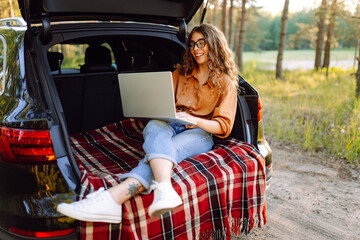 Young woman using laptop sitting in car trunk during sunset. Working while on vacation by car.