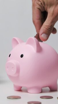 Vertical video. Hand places a coin in a piggy bank on a table amid scattered coins, signifying savings