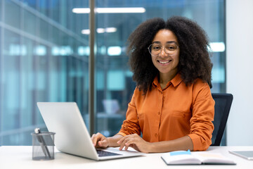 Smiling young professional woman working on a laptop at a modern office desk, exuding confidence...