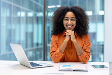 Smiling woman in bright office leaning on desk with laptop and open book, showcasing professional...