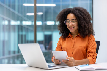 Smiling professional woman in orange shirt using digital tablet at desk with laptop in modern...