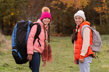 family walking in autumn forest with backpacks, girl in pink jacket, mother in orange sleeveless jacket, family in hat with pompoms, gen z