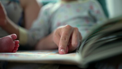 Close-up of mother pointing to a book while holding a baby and sitting with her older child on the couch, creating a family bonding moment filled with storytelling