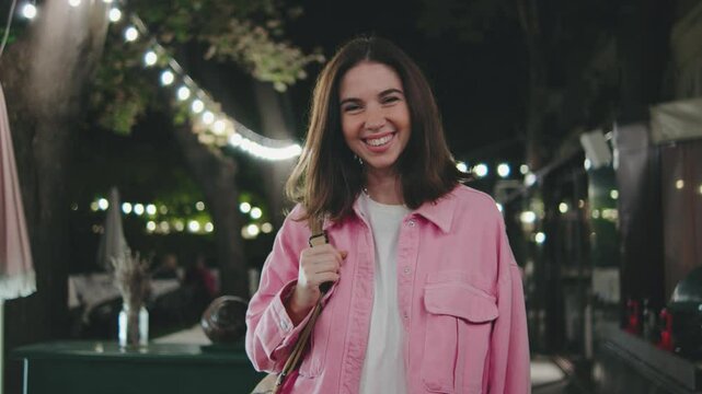 A joyful young woman is smiling brightly, surrounded by a festive evening setting filled with twinkling lights, creating an enchanting atmosphere perfect for celebration