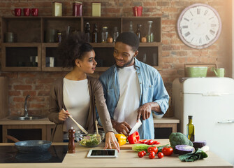 Happy african-american couple cooking healthy food and having fun together in their loft kitchen at home. Woman and man drinking wine. Preparing vegetable salad.