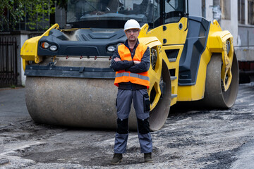 Construction worker stands confidently beside a yellow steamroller on a city street during daytime...