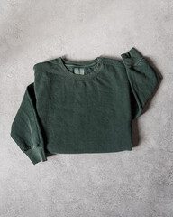 Blank Forest Green Crewneck Sweatshirt Folded Concrete Background, Stylized Sleeves, Natural Lighting with Bold Shadows, Simple Product Shot with Blank Tag, Casual Apparel Photography Mockup Template