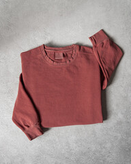 Blank Brick Red Crewneck Sweatshirt Folded on Concrete Background, Stylized Sleeves, Natural Lighting with Bold Shadows, Simple Product Shot with Blank Tag, Casual Apparel Photography Mockup Template