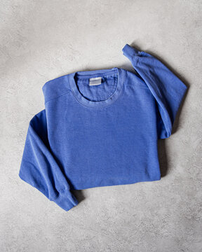 Blank Purple Blue Crewneck Sweatshirt Folded Concrete Background, Stylized Sleeves, Natural Lighting with Bold Shadows, Simple Product Shot with Blank Tag, Casual Apparel Photography Mockup Template