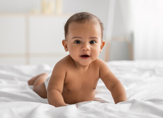 Portrait of cute small African American baby crawling on bed on the white blanket at home. Naked...