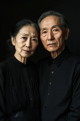 Obraz premium An elderly Asian couple poses closely in elegant black outfits. Their serious expressions against a dark backdrop emphasize their deep connection and timeless sophistication, making the image both