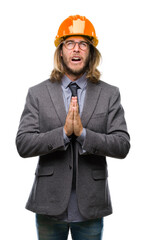 Young handsome architec man with long hair wearing safety helmet over isolated background begging and praying with hands together with hope expression on face very emotional and worried.