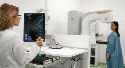 Female doctor examines breast scan on monitor while patient in blue gown undergoes mammogram...