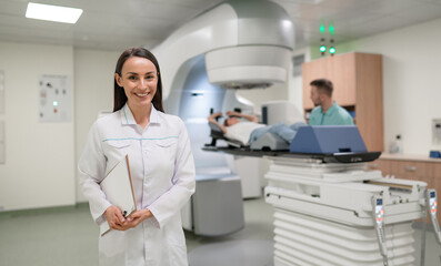 Smiling female doctor stands with clipboard in front of linear accelerator in modern medical...