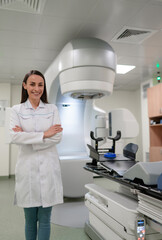 Naklejka premium Smiling female medical professional stands confidently in front of advanced radiotherapy equipment in modern clinic, wearing white lab coat. The image portrays a clean, high-tech healthcare setting