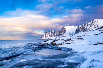 Tungeneset or Devils Jaw mountain on Senja island, Norway in winter. Winter landscape with snowy mountain peaks, fjord waters and ice coast in northern Norway. Scenic view with dramatic sky