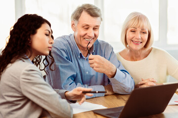 Beautiful Hispanic Travel Agent Working With Clients In Office, Offering Tours To Happy Elderly Couple