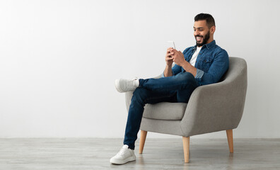 Joyful young Arab guy sitting in armchair with smartphone, working remotely or video chatting against white studio wall, empty space. Middle Eastern man watching movie, having online meeting