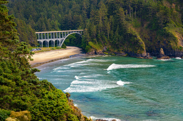 An arched bridge on the Oregon Coast Highway near Heceta Head Lighthouse, surrounded by forest, overlooking a sandy beach and turquoise ocean waters.