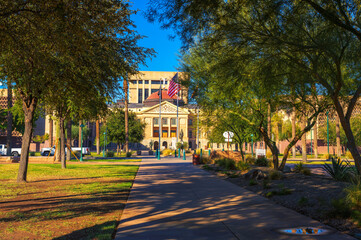 Naklejka premium Arizona State Capitol building in Phoenix, viewed from Wesley Bolin Memorial Plaza on at sunrise, with trees and flags.
