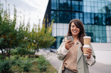 Businesswoman using smartphone and holding coffee walking outside modern office building