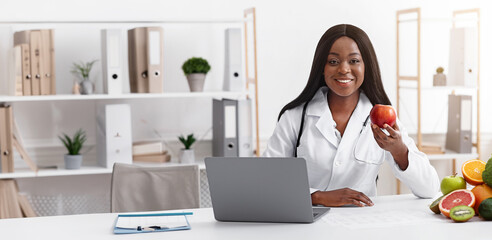 Pretty smiling black woman doctor nutritionist sitting at workplace in front of laptop and holding red apple, free space