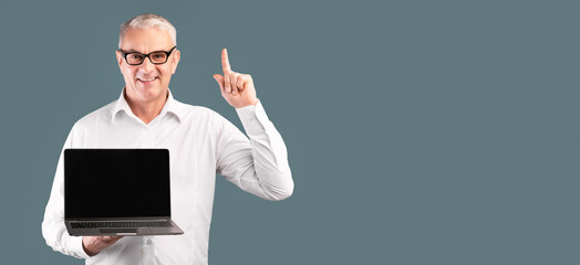 Useful Website. Middle-aged man pointing up and holding laptop with blank black screen over blue...