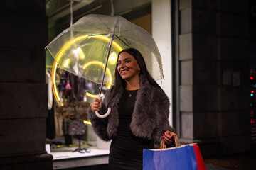 Young latin girl smiling while holding transparent umbrella as she shops in the city on rainy night