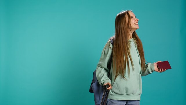 Joyous young traveler holding passport, going on vacation, looking around after arriving in new place. Happy girl travelling, sightseeing, visiting attractions, studio background, camera B