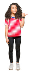 Young beautiful woman with curly hair wearing stripes t-shirt smiling with happy face looking and pointing to the side with thumb up.