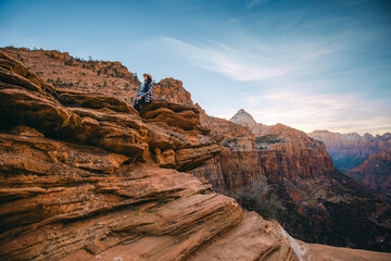 Woman in a hat and plaid jacket is sitting at a cliff of beautiful landscape view of Canyon Overlook point, one of the most favorite trail in Zion National Park, Utah, USA