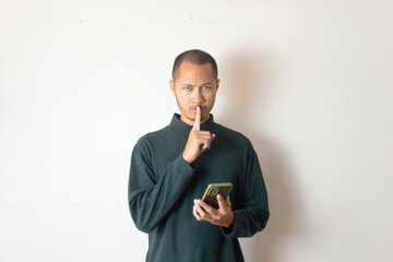 Young men wearing sweater silence gesture while holding phone isolated white