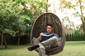 Young man reading a book relaxing in a comfy chair lounge in a green nature setting enjoying weekend holiday 