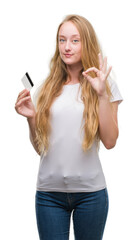 Obraz premium Blonde teenager woman holding credit card doing ok sign with fingers, excellent symbol