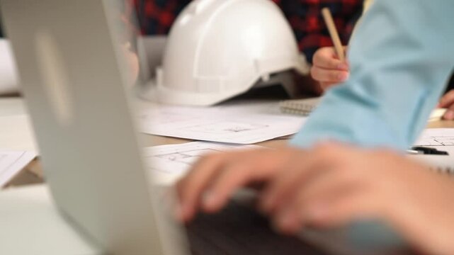 Architect and safety engineer discussion about building blueprint displaying on laptop on table with architectural document and wooden block scatter around at modern office. Closeup. Delineation.