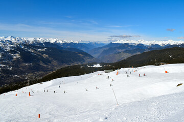 View of a mountain valley from the popular ski resort Meribel in France's Three Valleys. Skiers and snowboarders riding down a piste backdropped by breathtaking snowcapped French Alpine mountains.
