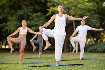 Sporty young adult man practicing yoga during group class in green peaceful summer park, performing one-legged pose on mat, focusing on balance, flexibility, and mental wellbeing