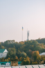 Obraz premium Scenic view of the Motherland Monument and a Ukrainian flagpole rising above a hilly autumn landscape with yellow and green trees, seen from a rooftop during a clear evening sky. 