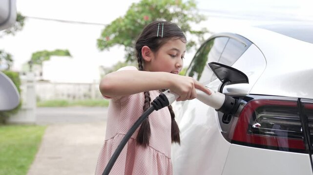 Happy little young girl learn about eco-friendly and energy sustainability as she recharge electric vehicle from home EV charging station. EV car and sustainable future generation concept. Fastidious