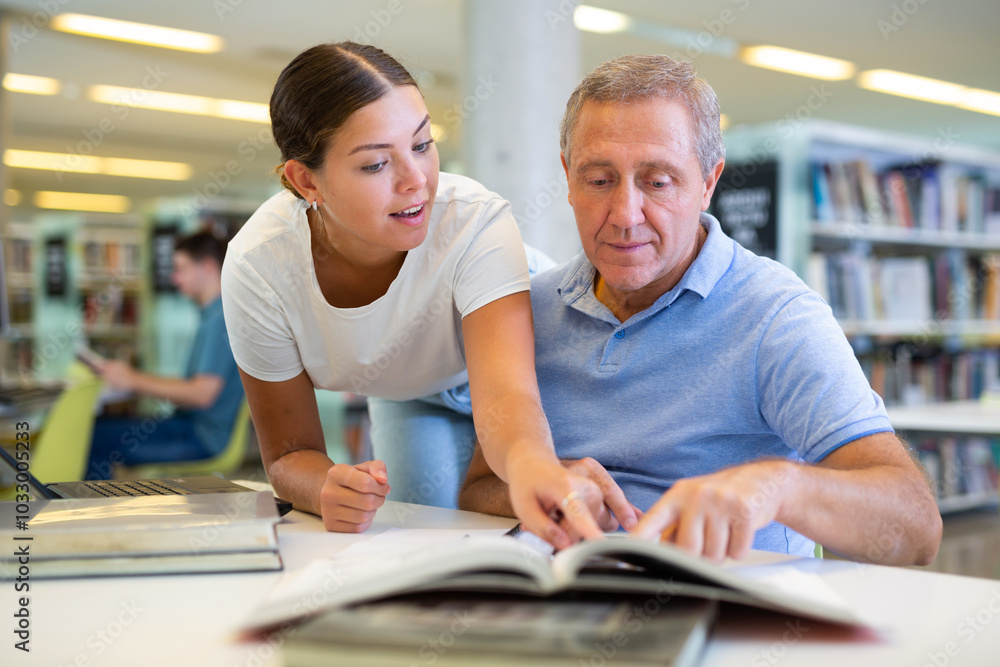 Poster positive young lady explaining information for mature man while he is using a computer in the librar