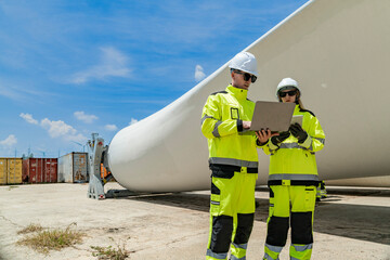 Two engineers in high visibility gear use a laptop to work in front of a large wind turbine blade at a construction site. the integration of technology in renewable energy and wind power projects.