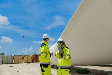 Two engineers in high visibility gear use a laptop to work in front of a large wind turbine blade at a construction site. the integration of technology in renewable energy and wind power projects.