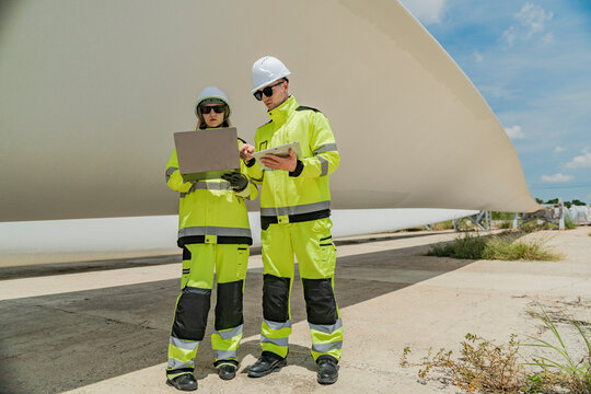 Two engineers inspect a massive wind turbine blade at a construction site. The image showcases the scale of wind power projects and the detailed work involved in renewable energy development.