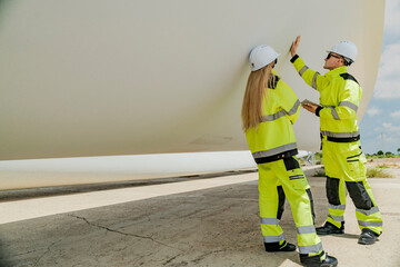 Two engineers inspect a massive wind turbine blade at a construction site. The image showcases the scale of wind power projects and the detailed work involved in renewable energy development.