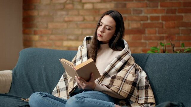 A woman sits comfortably on a couch, wrapped in a warm blanket, deeply engrossed in reading a book amidst a charming, cozy living space featuring a brick wall.