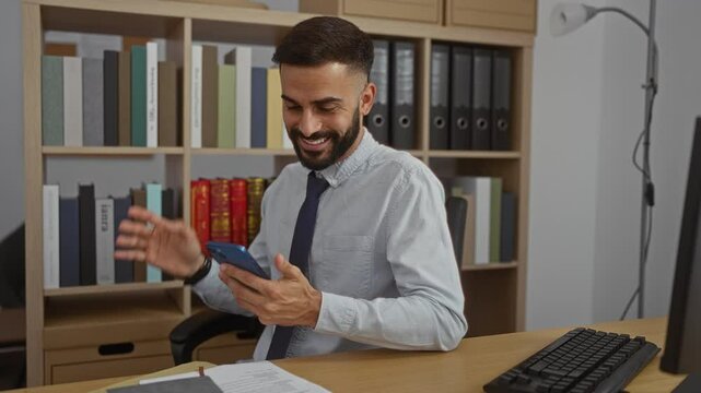 Young hispanic man with beard in office setting looking and smiling at smartphone for videocall with bookshelf and desk in the background, appearing professional and engaged.