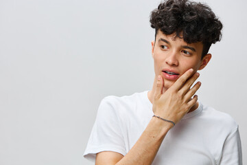 young man contemplating, touching his face, wearing a white t shirt, on a light grey background...