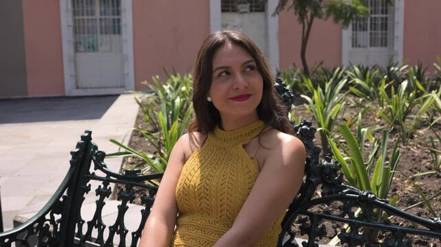 Mujer feliz mexicana y latina est&aacute; sentada en la banca de un parque y sonrie