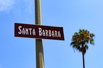 Fototapeta premium Santa Barbara sign is displayed on a pole, with a scenic backdrop of a blue sky and a tall, sunlit palm tree. Close up.